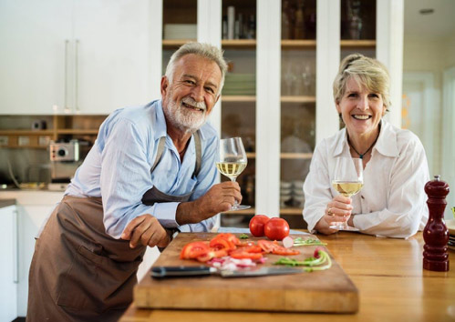 An image of an elderly couple in the kitchen, holding wine while they prepare food.