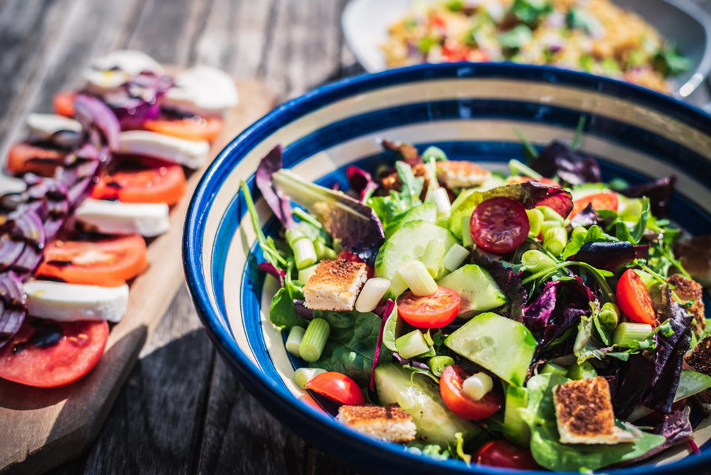 A blue ceramic bowl filled with a salad of leafy vegetables, tomatoes, and cucumbers, sitting atop a rustic table.